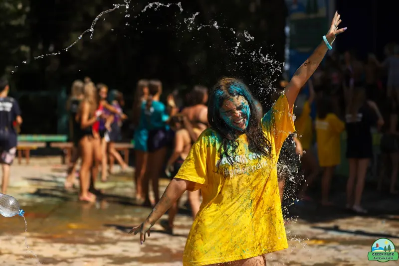 Festival-goer in a yellow T-shirt, face streaked with blue paint, arm raised as water sprays over the crowd