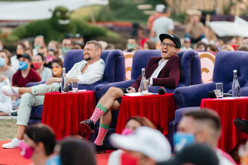 Guests in blue armchairs behind red tables on an outdoor stage, a masked audience in the foreground