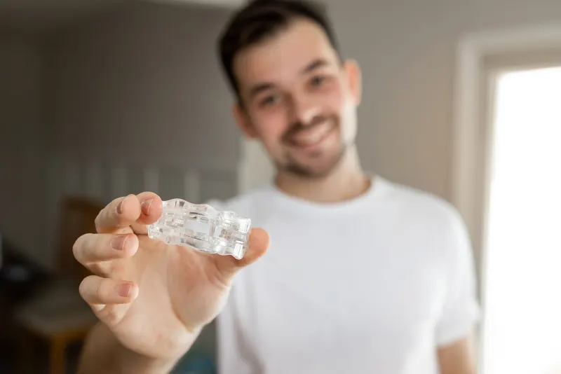 Smiling man holding a clear mouthguard towards the camera, the room behind softly blurred