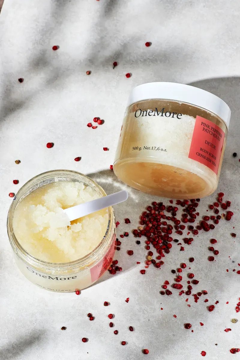 Two jars of body scrub on white stone scattered with red peppercorns