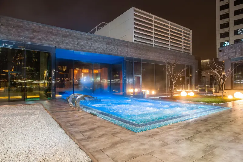 Lit pool terrace outside a glass building at night with a city block behind