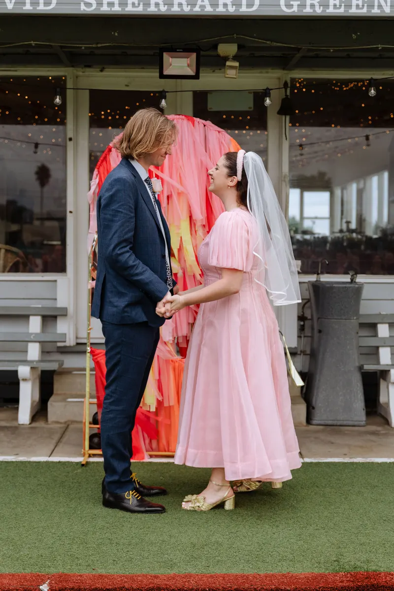 Bride in a pink dress and veil holding hands with the groom in front of a coral fringe backdrop