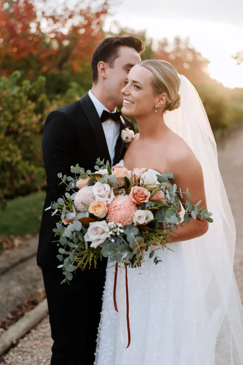 Groom kissing the bride's cheek as she holds a peach and cream bouquet, autumn trees behind