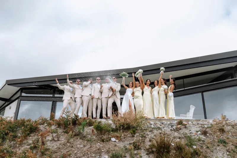 Wedding party in cream suits and champagne gowns cheering with bouquets raised in front of a glass-walled venue
