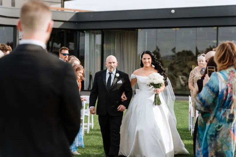 Bride walking the aisle on her father's arm past seated guests, glass venue behind