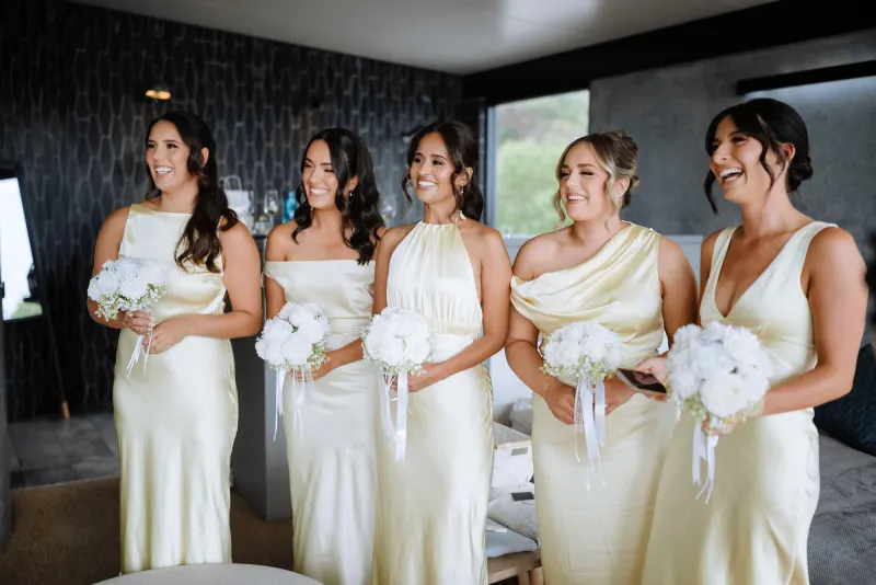 Five bridesmaids in champagne satin holding white bouquets against a dark textured wall