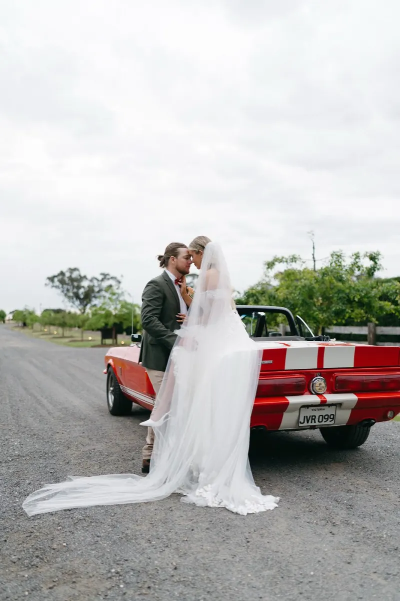 Bride and groom kissing beside a red convertible on a gravel road under an overcast sky