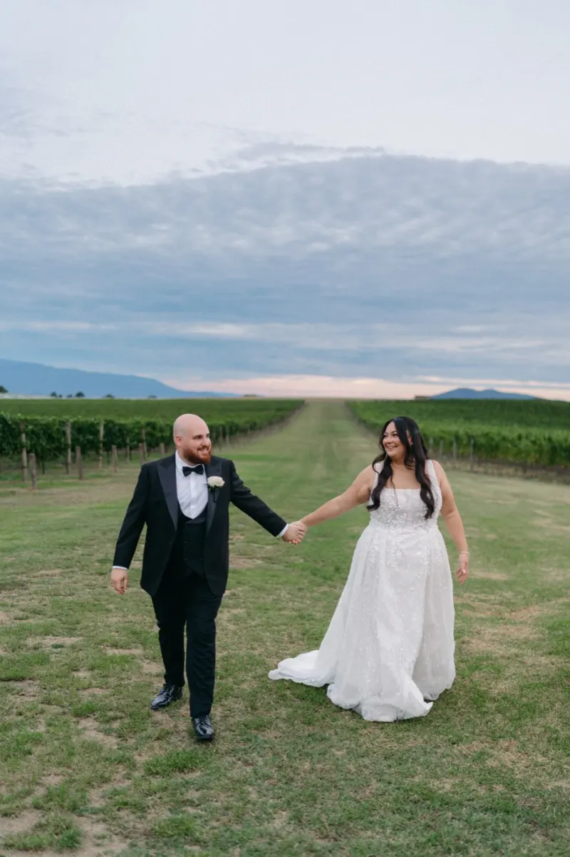 Bride and groom walking hand in hand through a vineyard under a streaked sky