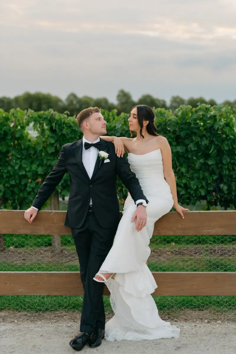Bride perched on a timber fence rail beside the groom in a vineyard